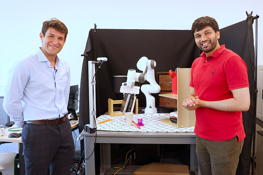 Marcel Torne Villasevil and Pulkit Agrawal stand in front of a robotic arm, which is picking up a cup