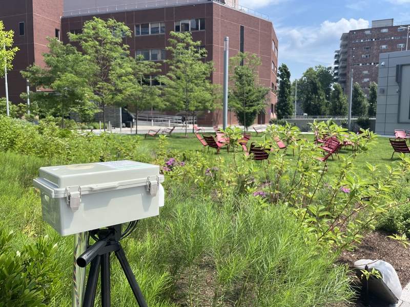 a tripod with small gray plastic box on top overlooks a lawn with chairs and buildings in the background