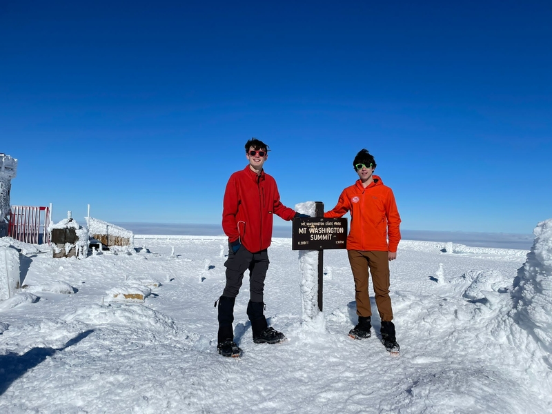 Nathanael Jenkins and Timur Uyumaz pose with the summit marker on Mt. Washington on a sunny day. They wear cold-weather gear and there is snow on the ground.