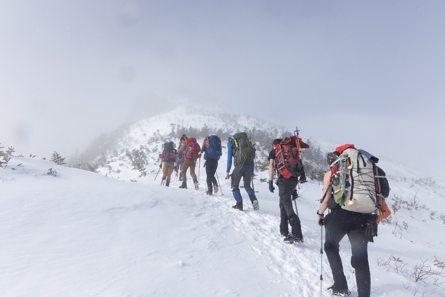 Seven students with large backpacks, photographed from behind, proceed in a line up a snow-covered slope.