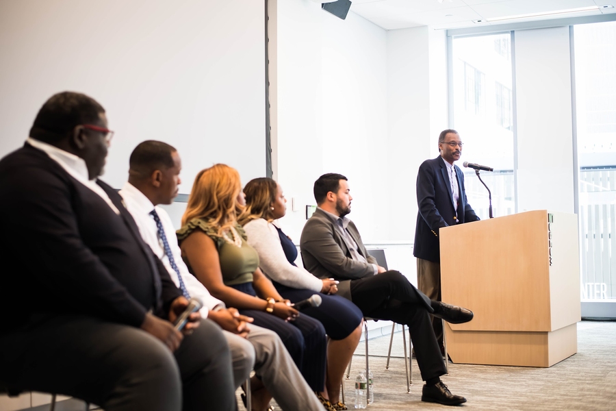 Five people seated on a stage watch a sixth person speak at a lectern