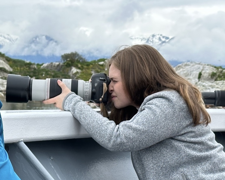 Side view of Sophie Hartley squinting into the viewfinder of a camera with a very large telephoto lens, with cloud-shrouded mountains in the background..