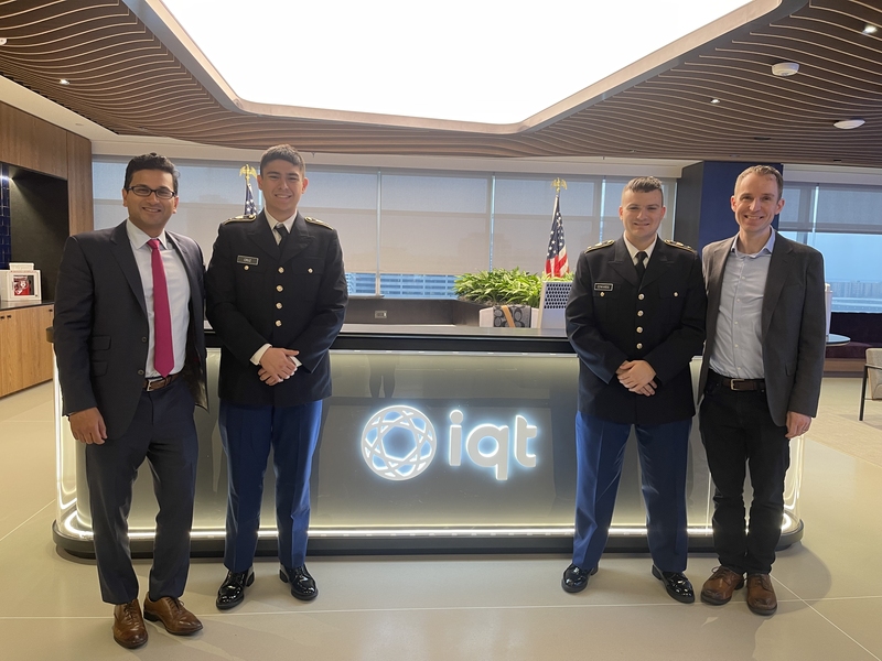 Aneal Krishnan, William Cruz, Alexander Edwards, and David LoBosco pose in front of a desk with a backlit “IQT” logo. Cruz and Edwards are wearing military uniforms.