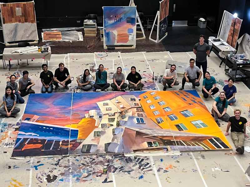 Sixteen individuals are kneeling or squatting around a large painted canvas depicting MIT's Stata Center, designed by architect Frank Gehry. The painted piece is colorful with bright orange and blue, showing the building at dusk. The piece is intended to be used for theater set.