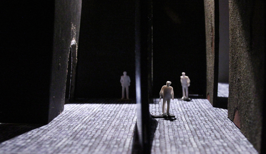 Close up shot of a physical model of a set design. White model silhouettes are walking on a paved surface, divided by walls.