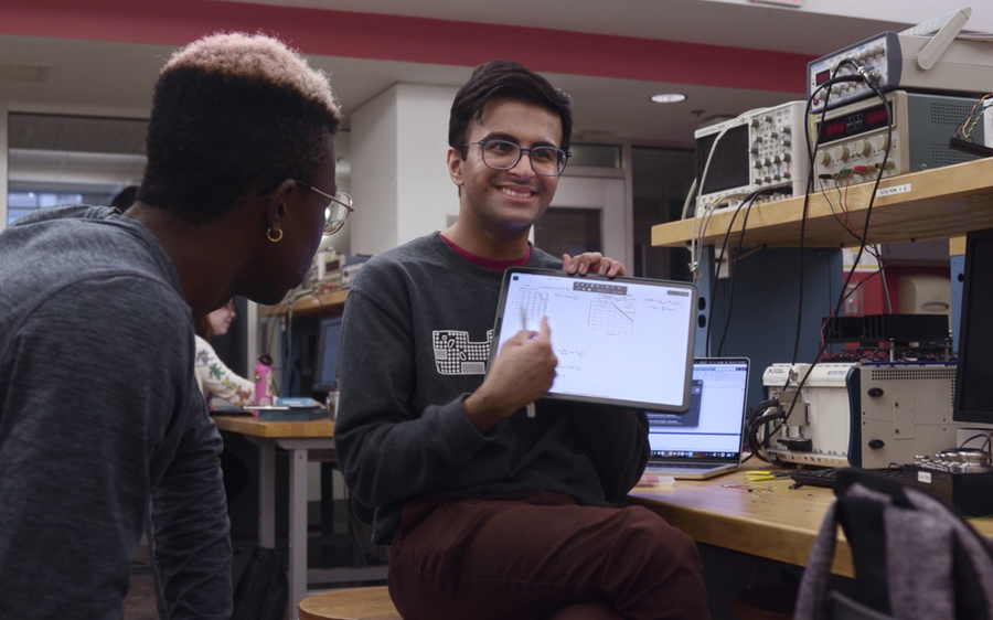 A student seated at a workbench points to a graph on a tablet computer facing the camera