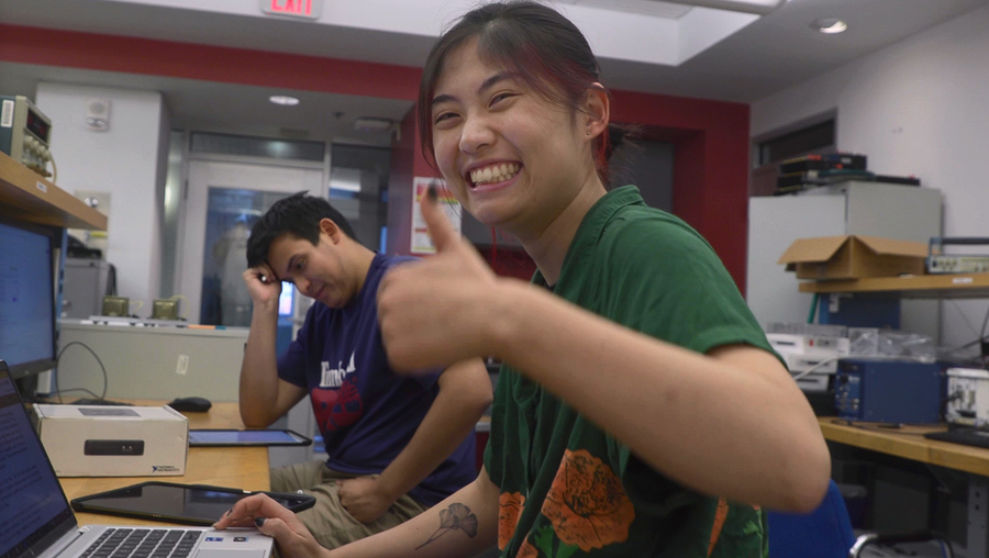 Audrey Cui sits at a laptop in a small lab, giving a thumbs up and a broad smile
