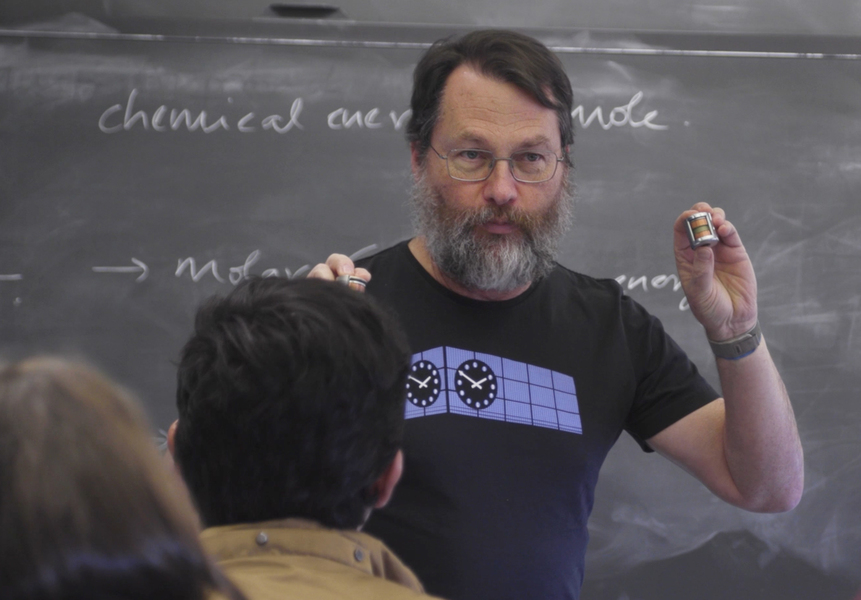 David Trumper stands in front of a chalkboard, holding up a small cylindrical electric motor in each hand