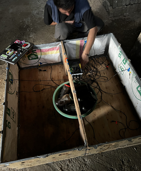 Ahmad Zakka squats by a wooden box that has been outfited with insulation and a bunch of wires