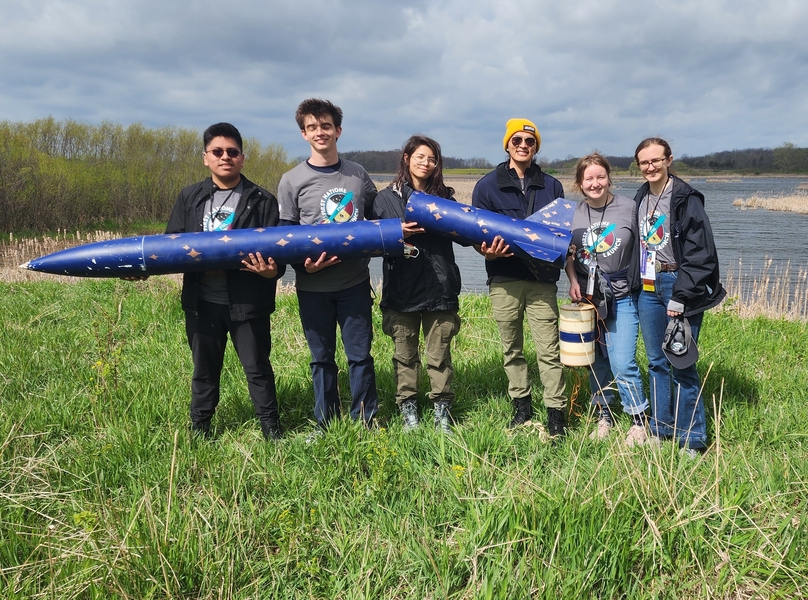 A group of six people stands together outdoors, holding a large blue rocket with gold star designs. They are dressed in team t-shirts and jackets, standing on green grass near a body of water.