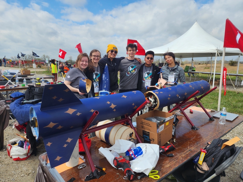 A group of six people stands together smiling at the camera behind a table covered with rocket components, tools, and equipment. The blue rocket with gold star designs is split into sections on the table, with parts being assembled.