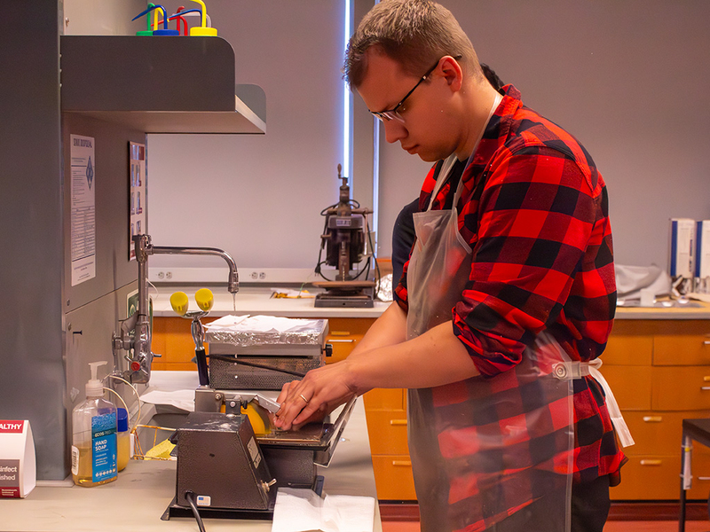 A summer course student wearing safety goggles and a clear plastic apron working at a benchtop saw