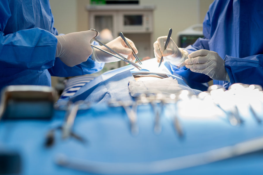 A zoomed in view of a surgery in progress. Blue gowned surgeons hold scalpels and tiny scissors above an opening in the patient's dressing where skin is exposed.