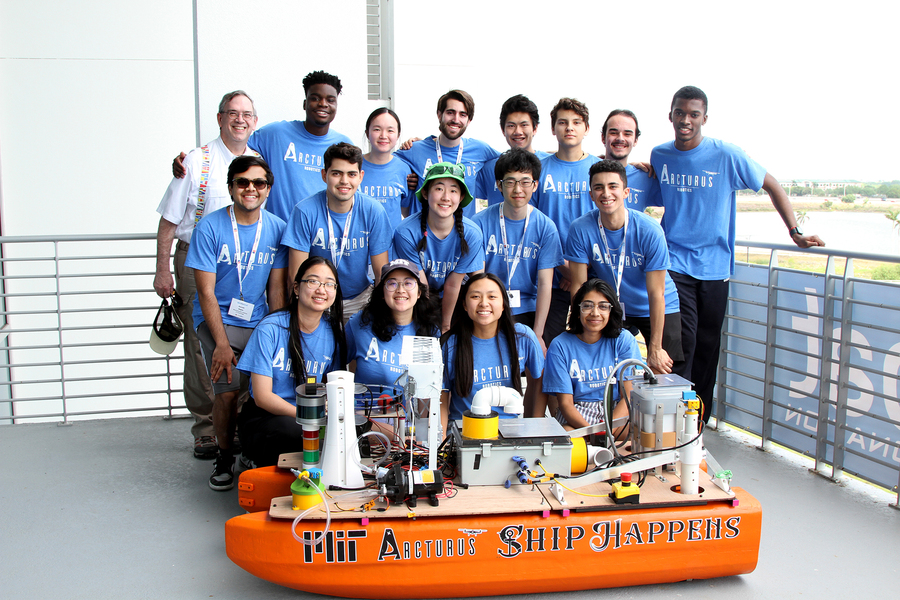 A group of 17 young people from the MIT Arcturus Robotics Team, wearing matching blue t-shirts, pose with their autonomous boat labeled "MIT Arcturus" and "Ship Happens" on a balcony overlooking a waterway, smiling proudly