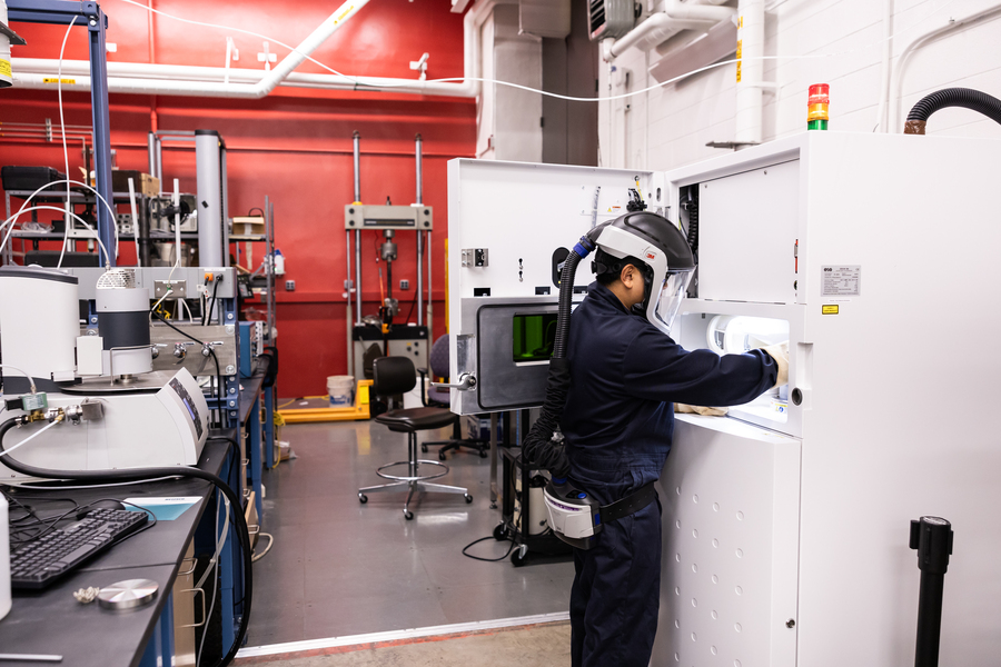 A student wearing protective gear including a full helmet and face shield with air supply works at a refrigerator-sized machine