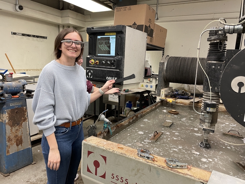 Hailey Polson wearing safety glasses holds up a machined part with another student behind her