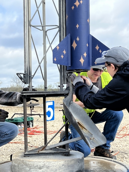 Two people work on securing a large blue rocket with gold star designs onto a launch pad. The people are both dressed in warm clothing. The rocket is positioned upright, and a large shovel head serves as a blast deflector.