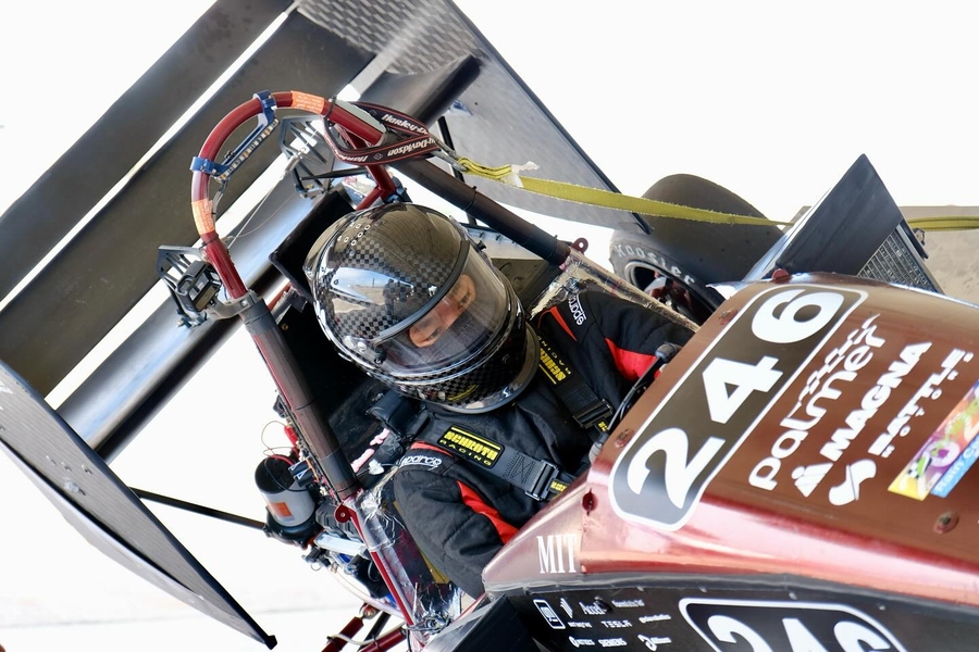 A student wearing a racing suit and helmet, sits focused in the cockpit of a Formula-style race car labeled "246," surrounded by the safety harness and roll cage, with the car's large rear wing and sponsor logos visible