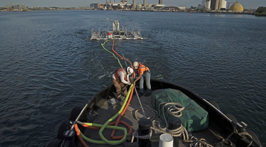 Two workers onboard a boat in Boston Harbor handle lines affixed to a barge-looking sonar array trailing behind the boat.