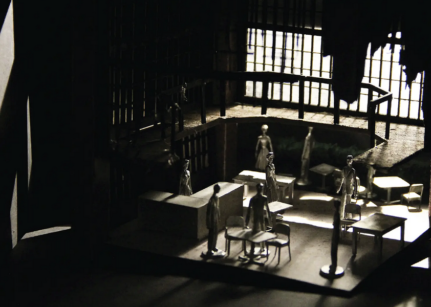 A back-lit set design model shows seven people standing on different floor levels near chairs and tables.