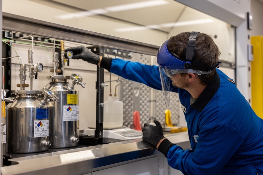 Zach Goodman, wearing full lab protection, adjusts a gauge above a stainless steel cylinder in a lab