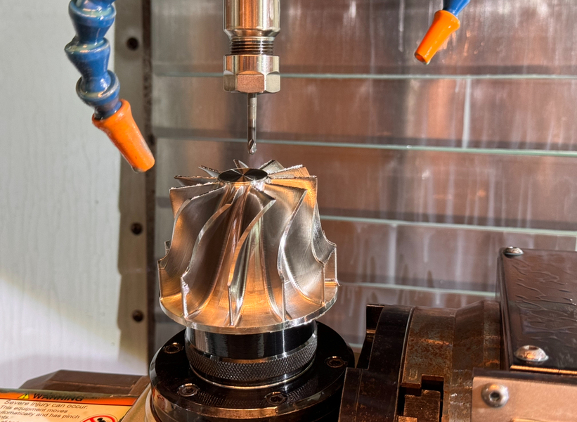 Close-up of a precision machining process shows a radial turbine with intricate curved blades being milled by a CNC machine, with a small cutting tool positioned above it and coolant nozzles aimed at the cutting area in a high-precision industrial setting.