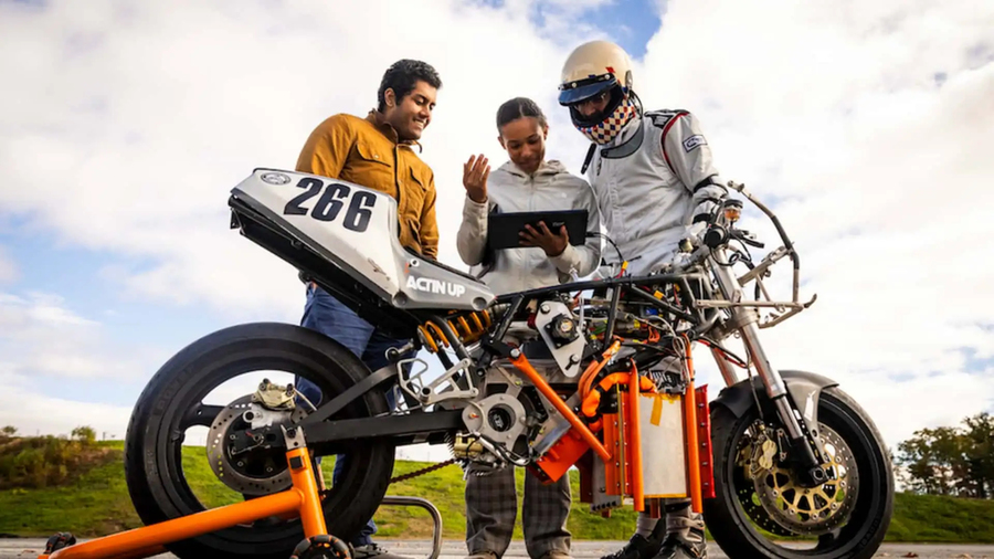 3 students gather around an orange motorcycle with a partially exposed design, showcasing intricate mechanical components