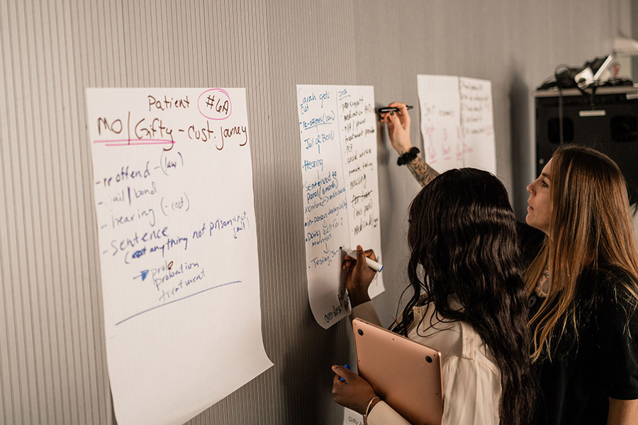 Two people write on one of several white notepads hung on a wall. 