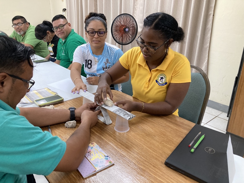 6 educators participate sit at a table. Three of them touch a small paper bridge holding a weight across its span.