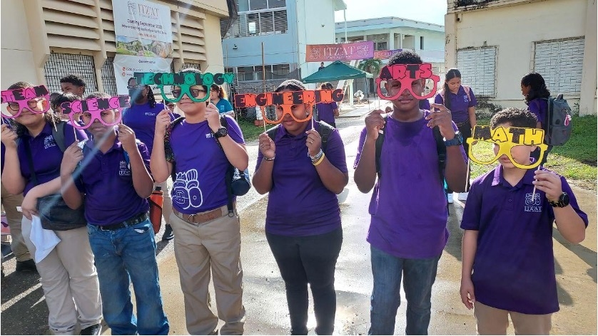 6 students wearing matching uniforms hold colorful glasses labeled with STEAM subjects, "Science," "Technology," "Engineering," "Arts," and "Math," in front of a school building