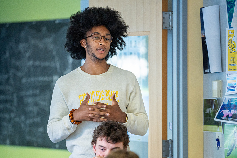 Will Bland stands and speaks before a blackboard with his fingers interlaced