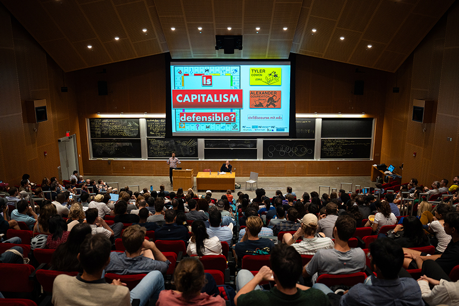 A full audience watches two men at the front of an auditorium-style classroom. A screen at front reads: "Is capitalism defensible?"