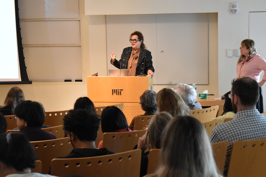 Erin Schuman stands and gestures behnind a wooden lectern with the MIT logo on it. Her audience is in the foreground, sitting in rows of auditorium-style chairs.