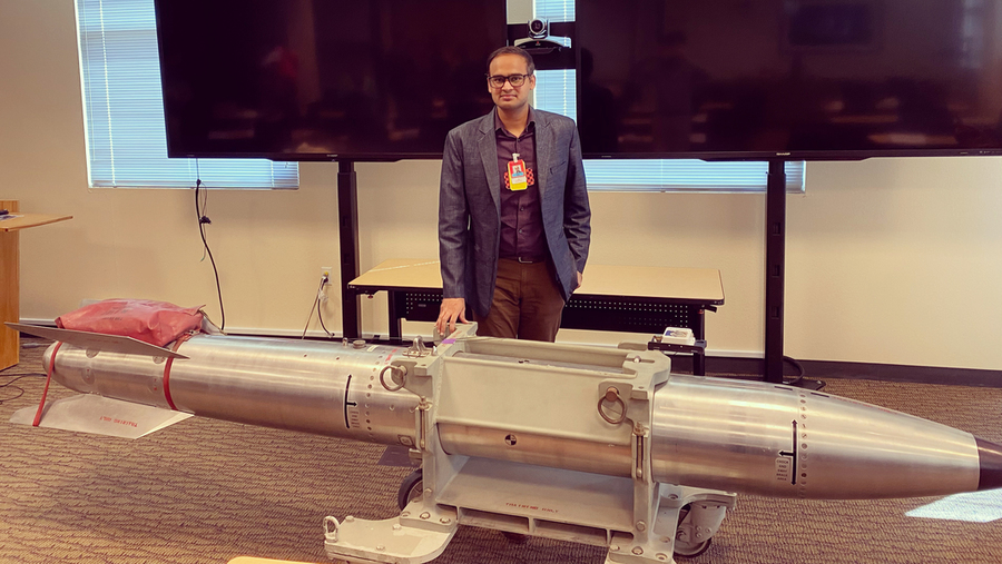 Kunal Singh stands before a silver missile in room with a flat screen behind him. The rocket looks to be about 3.5 meters long.
