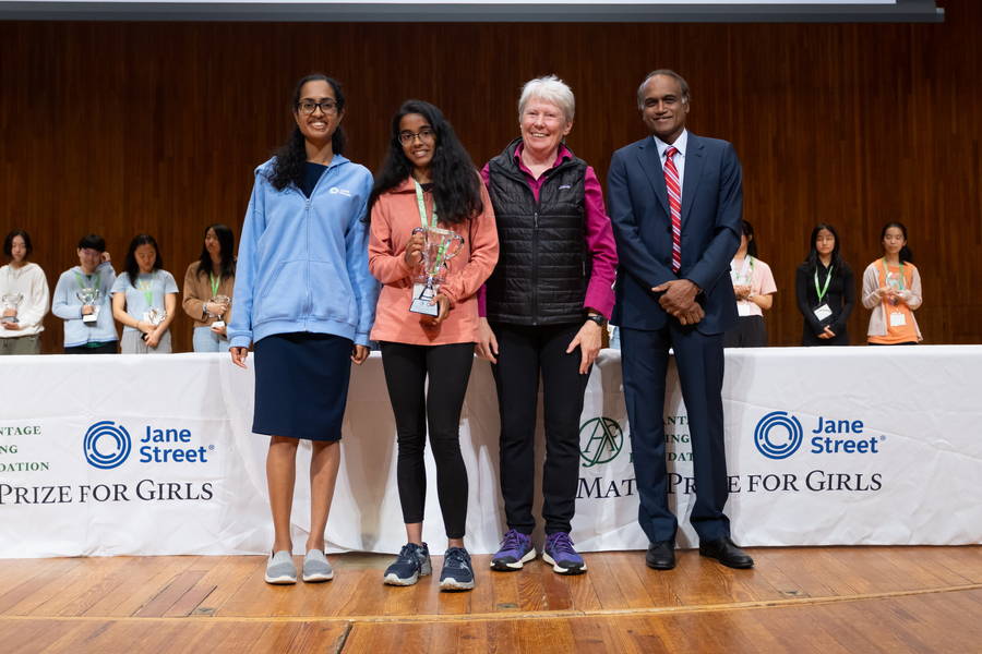 Nitya Mani, Shruti Arun, Maria Klawe, and Arun Alagappan pose together on a stage. Arun holds a trophy.