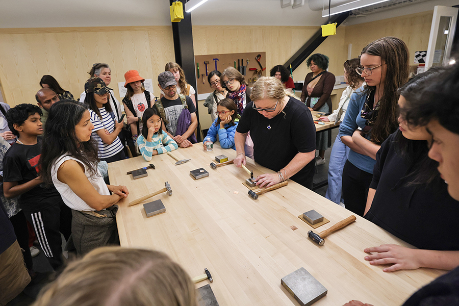 Lots of people stand around a table, which holds small square slabs of silver metal and some hammers