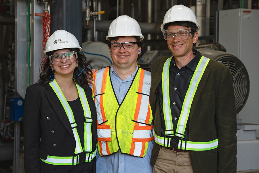 Shreya Dave, Brent Keller, and Jeffrey Grossman pose together in hard hats, safety goggles, and visibility vests in front of some industrial equipment