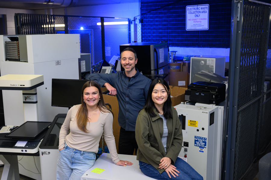 Anastasia Dunca, Chris Rabe, and Jasmin Liu stand in an electronic waste collection area, surrounded by electronic and lab equipment