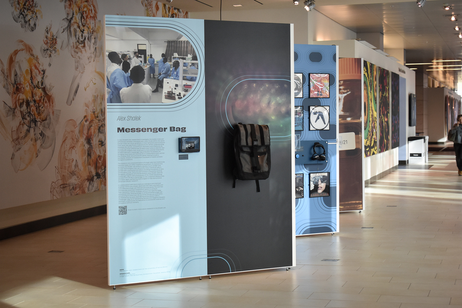 A exhibit panel in a gallery displaying a photo of numerous scientists in a lab as well as a small device and a large grey backpack