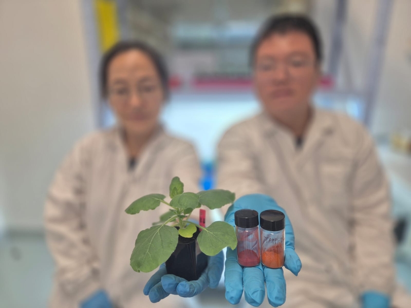 Two out-of-focus scientists in a lab. One holds up a small potted plant; the other, two small glass vials containing powder in different shades of red