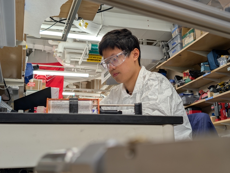 ZhiYi Liang, wearing protective gear, sits at a table with scientific equipment