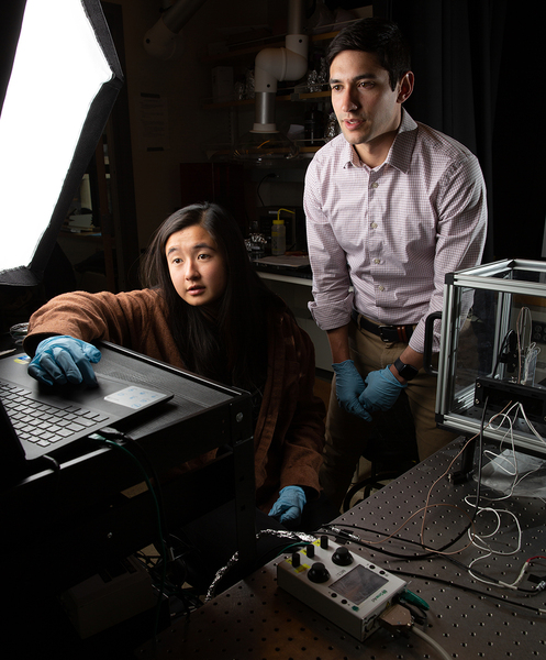 Carlos Portela and Rachel Sun, wearing nitrile gloves, look at a laptop screen in a darkened lab room.