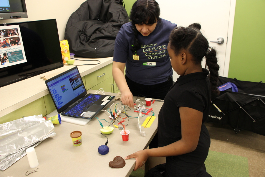 A woman helps a girl design a controller using playdough, an apple, a plastic cup, and tinfoil as the controller keys to play a piano on a computer.