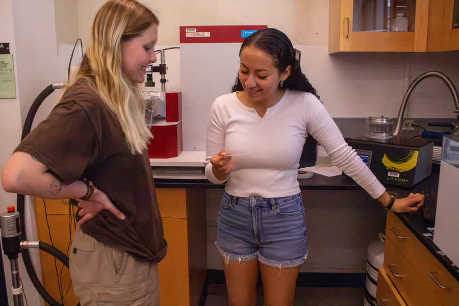 Victoria Dzieciol and Diane Batres stand in a laboratory looking at a glass slide and talking.