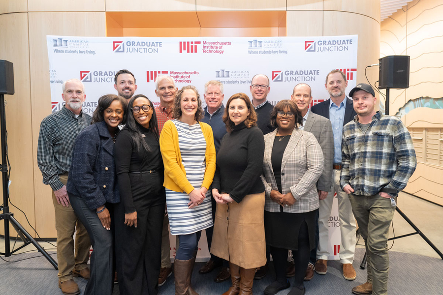 13 people pose standing in two rows before a poster that says "MIT" and "Graduate Junction"