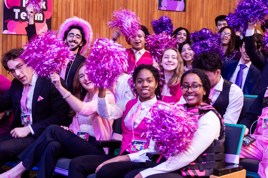 A group of people dressed in pink and with pom-poms cheer in the audience.