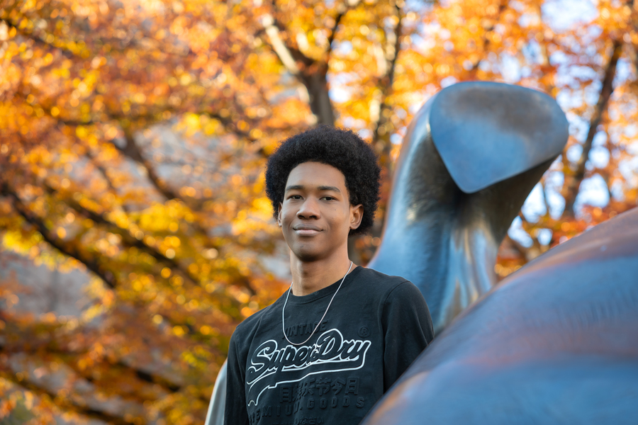 Ronaldo Lee stands cradled by an MIT statue, with yellow leaves on the tree behind him.