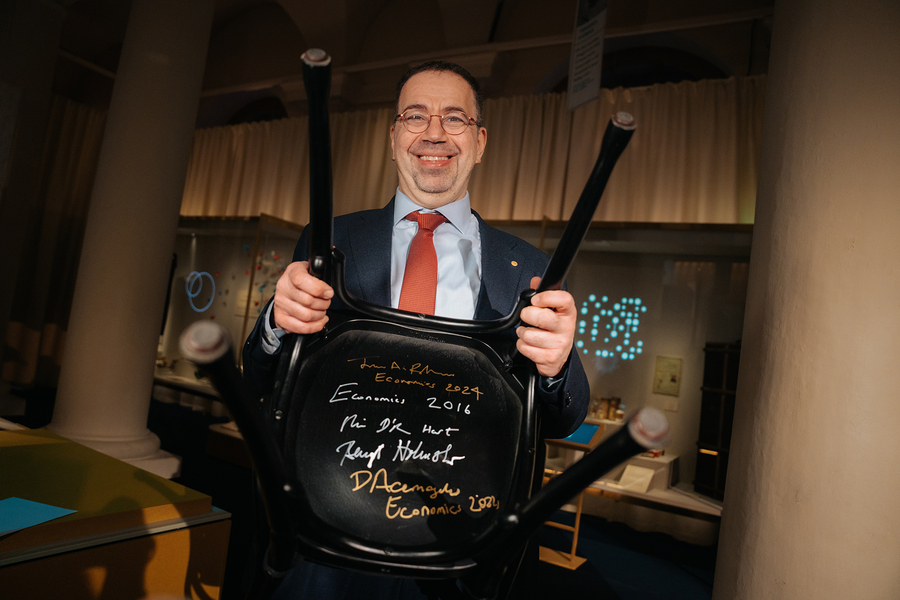 Daron Acemoglu holds up a black chair to show the bottom, which is signed with various Nobel laureates' signatures
