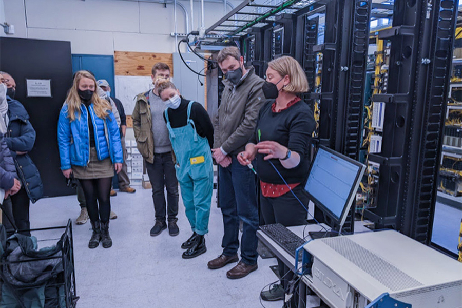 Seven people with face masks stand near a computer in a space filled with wired fiber optic data hub. One is speaking to the rest.