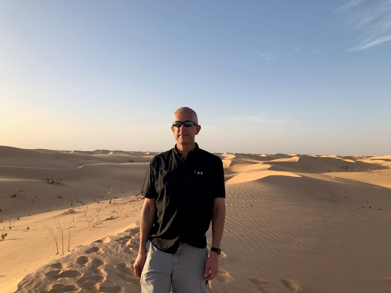 Matthew Donal stands in a desert with dunes behind him on a sunny day.
